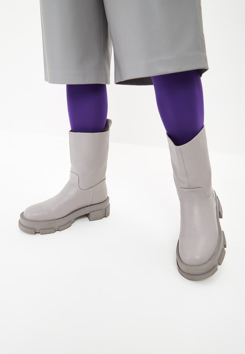 Botas de tobillo grises con un acabado de cuero suave, suela gruesa y texturizada, y un eje ancho. Llevadas con medias moradas y un atuendo gris.