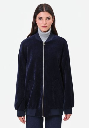 Donna con capelli lunghi e scuri indossa una giacca di velluto blu navy con zip sopra un dolcevita grigio, in piedi davanti a uno sfondo bianco uniforme.
