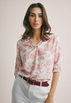 Kobieta o brązowych włosach do ramion, ubrana w białą bluzkę z różowymi kwiatowymi wzorami oraz białe spodnie z ciemnoczerwonym paskiem.