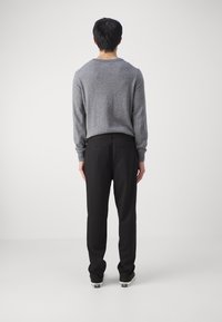 Mężczyzna stojący tyłem w szarym sweterze, czarnych spodniach i czarno-białych trampkach na jednolitym białym tle.