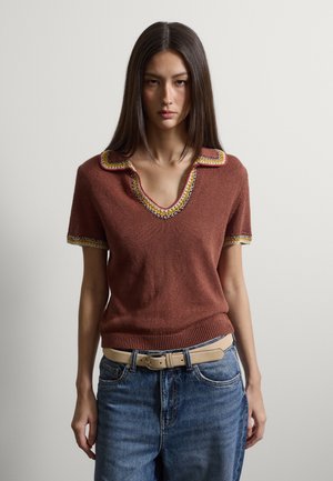 Mujer con cabello largo y oscuro, vestida con una blusa de punto de manga corta color óxido con ribetes multicolor y jeans azules con cinturón beige, de pie contra una pared blanca.