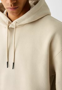 Man som bär en beige hoodie med dragsnodd och svarta spetsar, synlig från axlarna till läpparna.