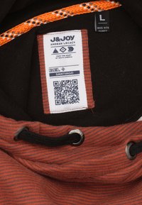 Zwarte capuchon kledingstuk met een oranje gestreepte trekkoord, een gestructureerde roestkleurige stof, en een label met QR-code en merkinformatie.