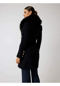 Abrigo de lana negro con cuello de piel, con botones dorados en las mangas y una silueta entallada. Se combina con pantalones azul oscuro.