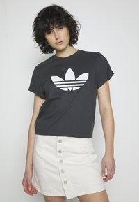 Tamsiai pilka medvilnės marškinėliai su baltu Adidas logotipu, kuris turi tris lapus ir juostas, derinami su balta džinsine sijonu su mygtukais.