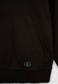 Ciemnobrązowa bluza z kapturem z kieszenią z przodu, ściągaczami na mankietach i u dołu. Posiada czarną metkę z białym tekstem oraz akcentami graficznymi w pobliżu kieszeni.