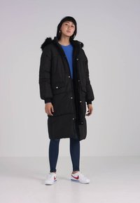 Manteau long noir en duvet avec capuche en fourrure, doté de boutons-pression, de poches avant et d'une texture matelassée. Porté par-dessus un haut bleu et un jean.