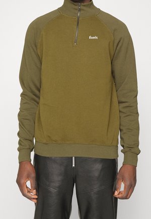 Olivgrüner Quarter-Zip-Pullover mit gerippten Bündchen und Saum, getragen mit schwarzen Lederhosen, zeigt Vorderteil des Torsos und Arme.