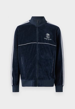 Teget plava velurova jakna na patent s bijelim prugama na rukavima i logotipom Tommy Hilfiger na lijevoj strani prsnog dijela, s elastičnim manšetama i rubom.