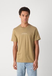 Khaki cotton t-shirt with short sleeves, round neckline, and white Calvin Klein Jeans logo across the chest. Paired with light blue jeans.