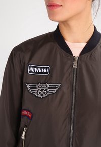 Giubbotto bomber marrone con colletto a coste nero, chiusura con zip e tre toppe ricamate che rappresentano "NOWHERE" e "ROUTE 66."