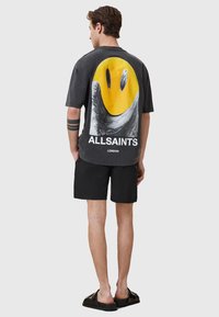 T-shirt nera oversize con una grande grafica di faccina sorridente gialla, accenti bianchi e grigi, e il testo "ALLSAINTS LONDON" in basso.