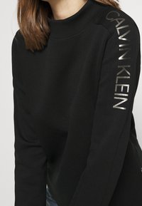 En person med brunt hår bär en svart sweatshirt med "CALVIN KLEIN" tryckt i silver på högra ärmen, poserande mot en neutral bakgrund.