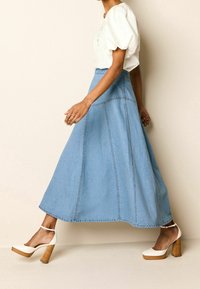 Jupe maxi en denim bleu clair avec des détails de couture ; associée à des chaussures à talons hauts blanches à bride de cheville et un blouse blanche à manches courtes.