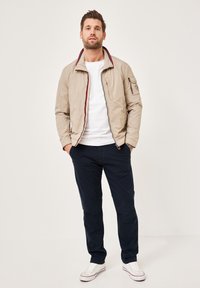 Beige Bomberjacke mit einem rot-blauen Kragen, mit Reißverschluss und seitlichen Taschen, kombiniert mit blauen Hosen und weißen Sneakers.