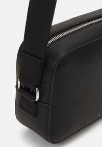 Borsa rettangolare nera realizzata in materiale liscio, con una zip argentata, tracolla regolabile e un attacco per la tracolla testurizzato.