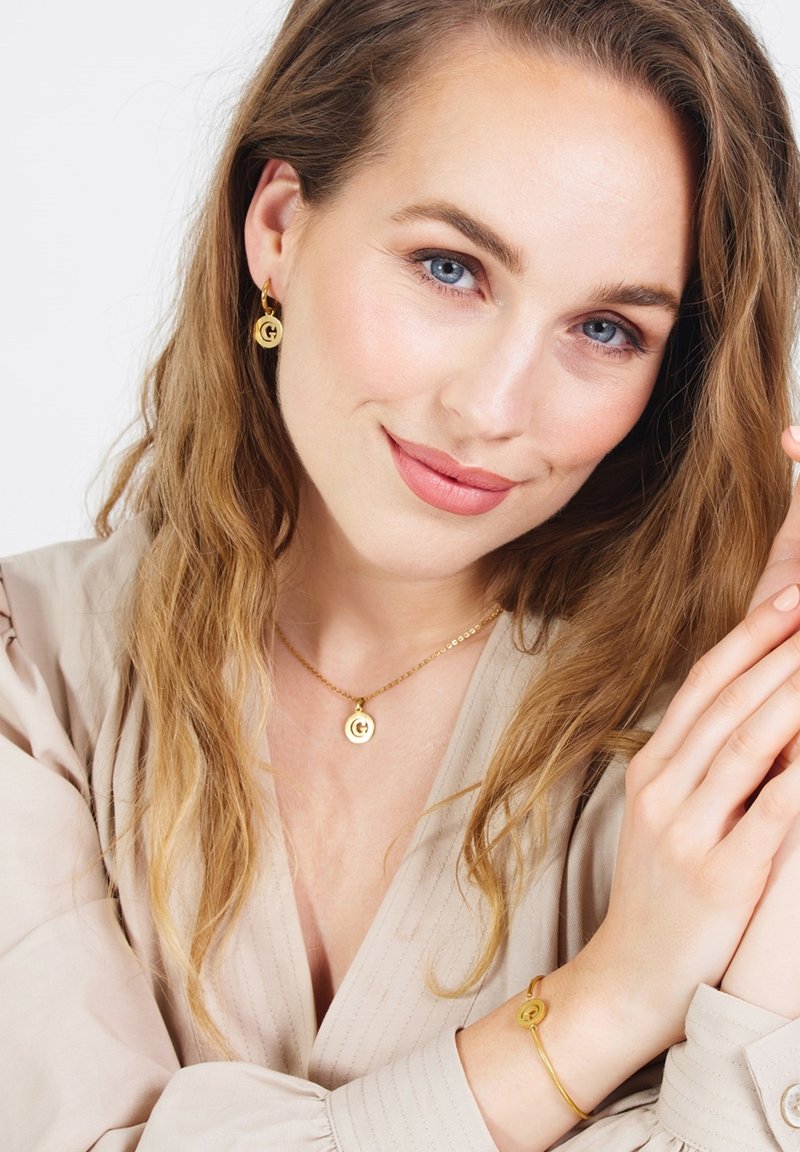 Gouden sieradenset met ronde oorbellen met logo, een bijpassende ketting met een ronde hanger en een delicate armband. Gladde textuur.