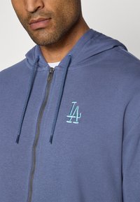 Blå zip-hoodie i stof med kengurulomme, justerbar hætte og lyseblå broderet logo på venstre bryst.
