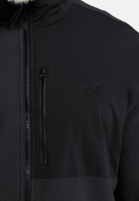 Giacca nera con taschino sul petto con cerniera, un mix di tessuto liscio e strutturato, e logo in rilievo sul lato sinistro superiore.