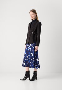 Svart blazer med detalj i sølvring, kombinert med en blå og hvit mønstret skjørt. Modellen har på seg svarte ankelstøvler. Jevne teksturer.