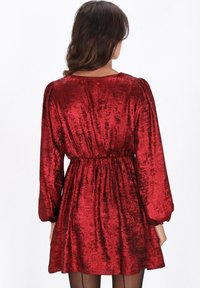 Vestido rojo con textura metálica, mangas largas y cintura ceñida. Presenta una falda acampanada y mangas abullonadas, creando una silueta voluminosa.