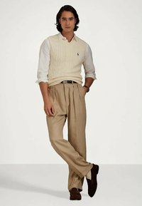 Jeune homme debout, portant une chemise blanche avec un gilet en tricot crème, un pantalon beige plissé, des chaussures marron foncé et une montre au poignet.