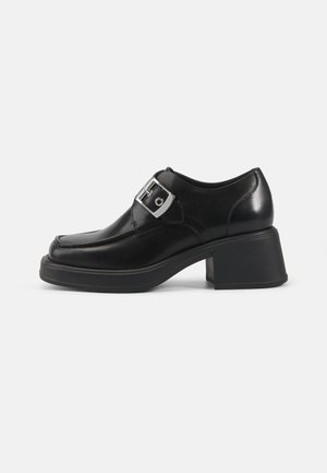 Black leather shoe with a rounded toe, block heel, and a side buckle detail. Smooth texture with minimalistic design elements.