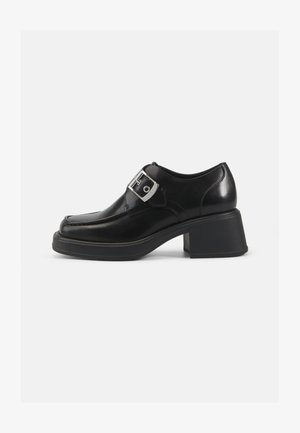 Chaussure en cuir noir avec un orteil arrondi, un talon bloc et un détail de boucle sur le côté. Texture lisse avec des éléments de design minimalistes.