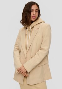 Beige pinstriped blazer with a loose fit, featuring wide lapels, a single button closure, and a pocket; layered over a beige hoodie.