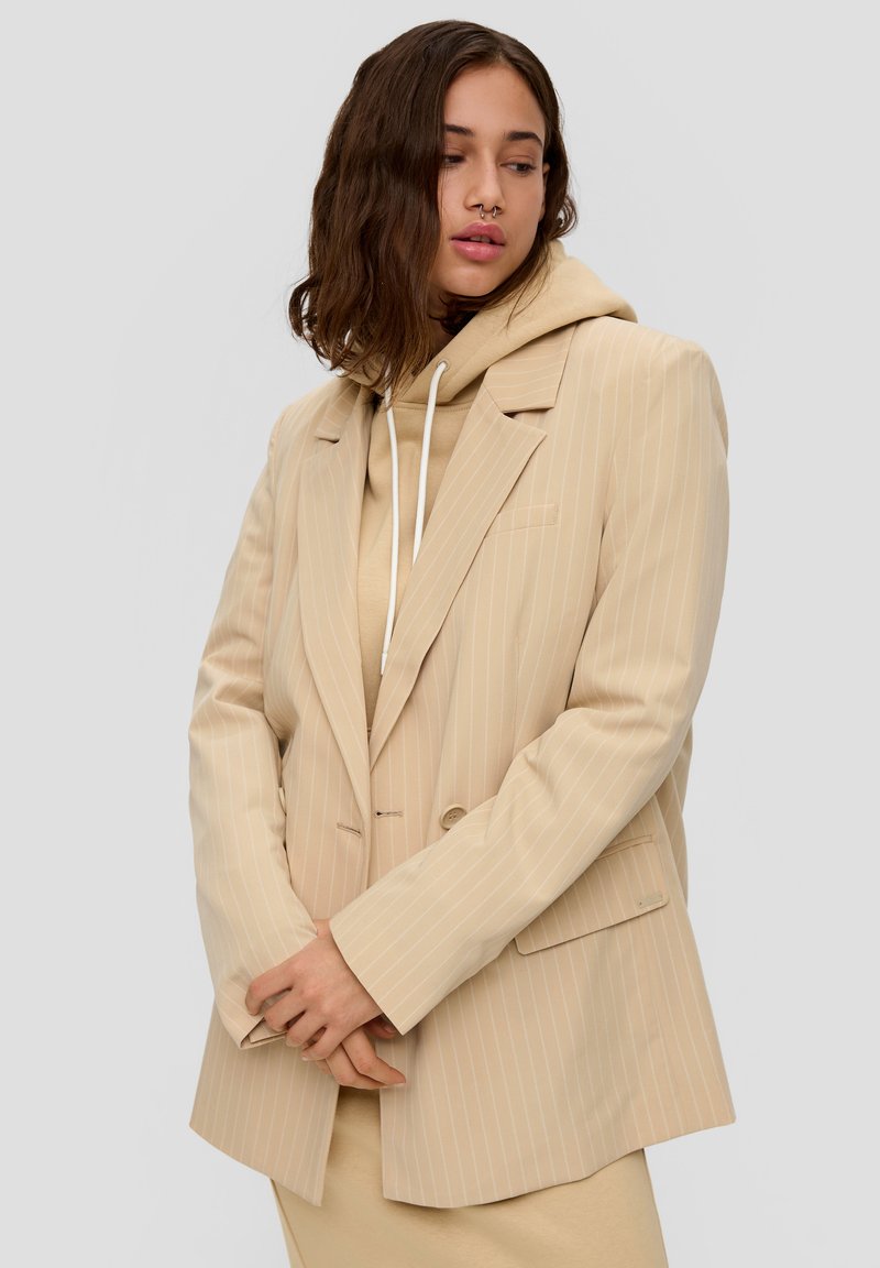 Beige pinstriped blazer with a loose fit, featuring wide lapels, a single button closure, and a pocket; layered over a beige hoodie.