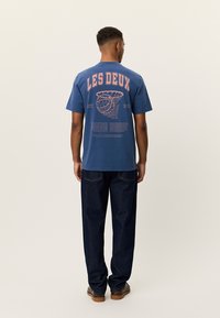 Μπλε βαμβακερό μπλουζάκι με μεγάλο πορτοκαλί τύπωμα "LES DEUX" και γραφιστικό σχέδιο στην πλάτη, συνδυασμένο με σκούρο τζιν παντελόνι και καφέ παπούτσια.