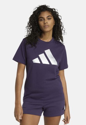 Joven mujer con cabello rizado que lleva una camiseta Adidas morada oscura con logotipo y pantalones cortos a juego contra un fondo liso y claro.