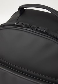 Schwarzer Rucksack mit einer glatten, matten Oberfläche. Verfügt über einen stabilen Griff, doppelte Reißverschlüsse und dezente Nähte entlang der Kanten. Kompakte Bauweise.