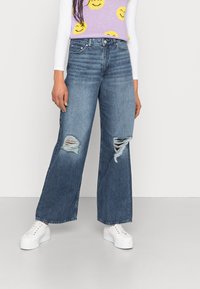 Jeans de perna larga em denim azul médio, com rasgos desgastados nos joelhos, combinados com uma blusa branca e ténis brancos de plataforma.