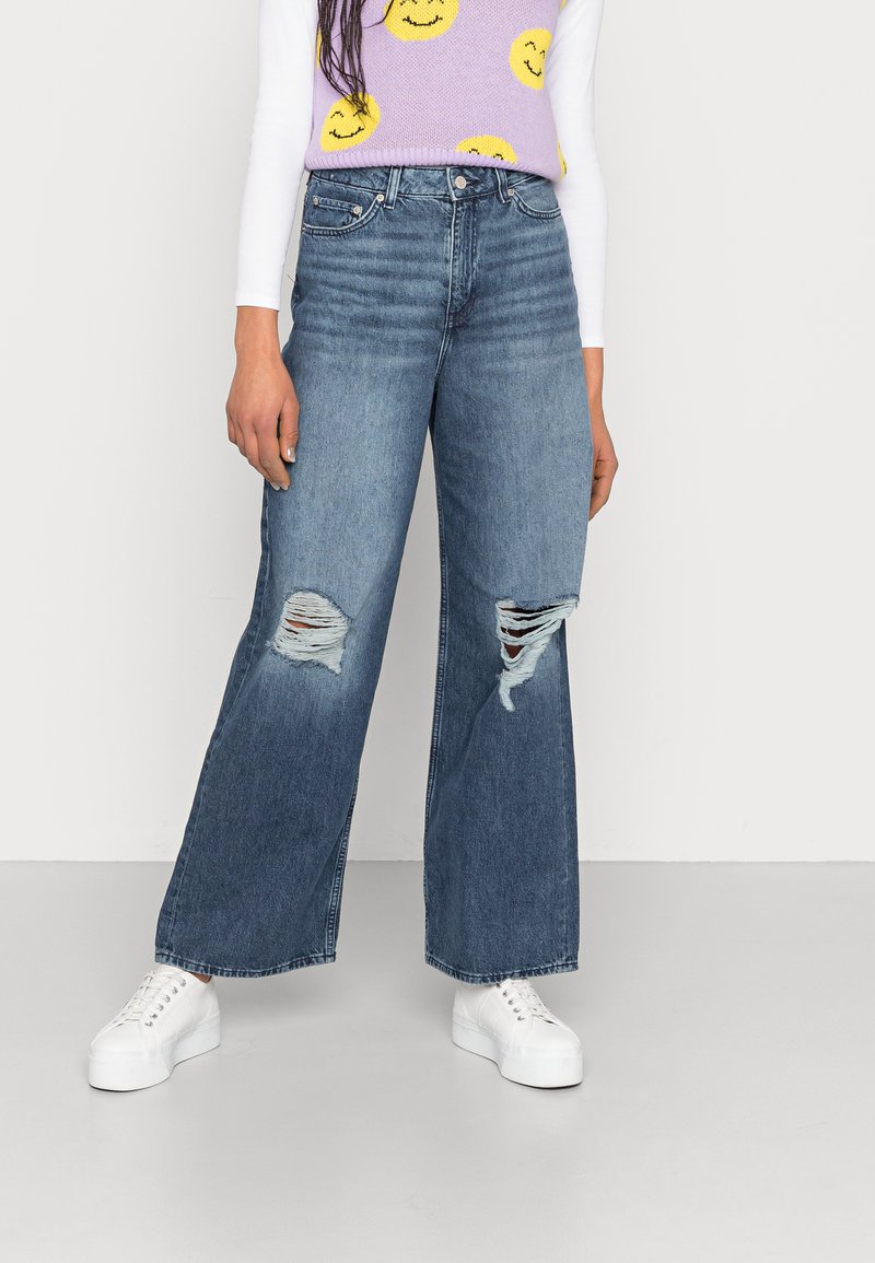 Jeans de perna larga em denim azul médio, com rasgos desgastados nos joelhos, combinados com uma blusa branca e ténis brancos de plataforma.