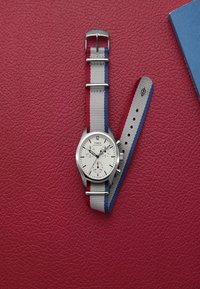 Silberne Uhr mit weißem Zifferblatt, ausgestattet mit drei Subdials und einem grauen Stoffarmband mit roten und blauen Streifen, platziert auf einem strukturierten roten Hintergrund.