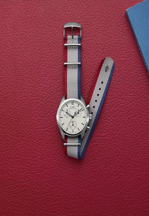 Silberne Uhr mit weißem Zifferblatt, ausgestattet mit drei Subdials und einem grauen Stoffarmband mit roten und blauen Streifen, platziert auf einem strukturierten roten Hintergrund.