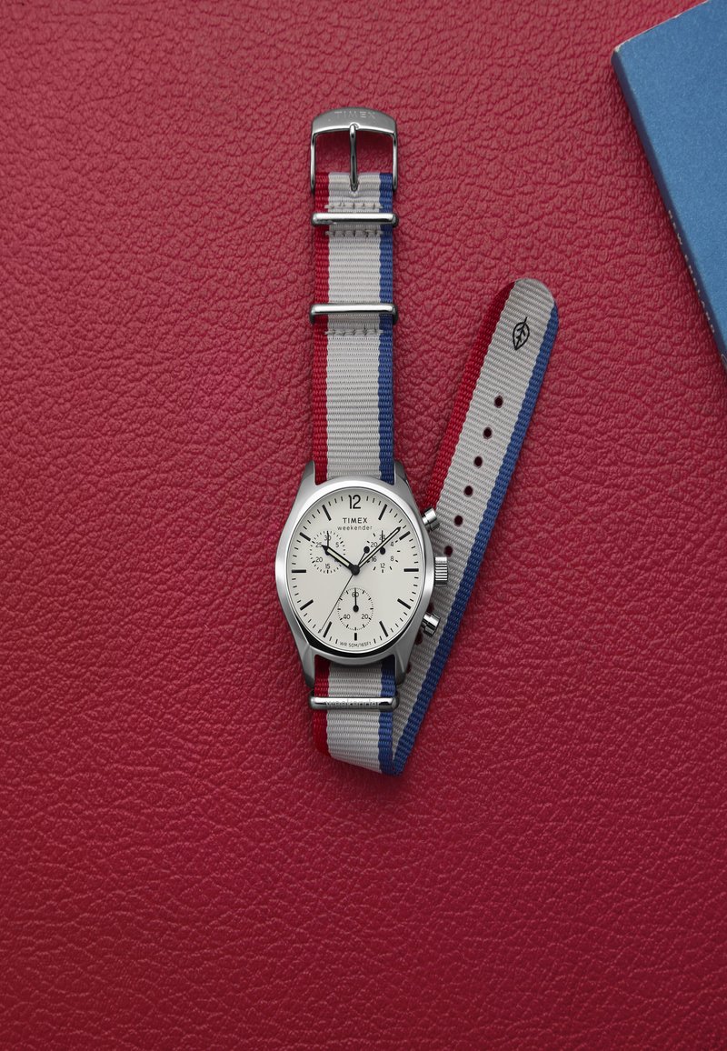 Silberne Uhr mit weißem Zifferblatt, ausgestattet mit drei Subdials und einem grauen Stoffarmband mit roten und blauen Streifen, platziert auf einem strukturierten roten Hintergrund.