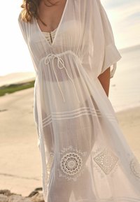 Witte, vloeiende kaftan van lichtgewicht, doorschijnende stof. Heeft een strik taille, borduursel aan de onderrand en ontspannen, brede mouwen.