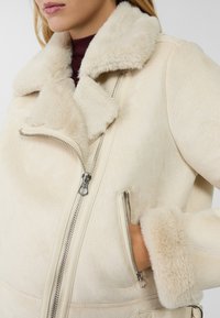 Béžová bunda z umělé kožešiny se širokým límcem, zapínáním na zip vpředu a velkými bočními kapsami. Vyznačuje se měkkou strukturou a kontrastním lemováním kožešinou.