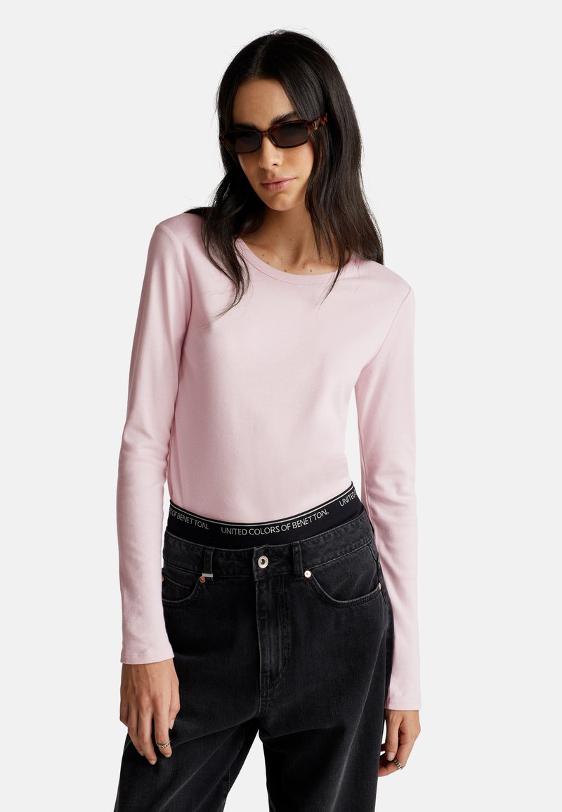 Kobieta nosząca okulary przeciwsłoneczne w muszki, jasnoróżową bluzkę z długim rękawem oraz czarne jeansy z wysokim stanem z elastycznym paskiem z logo.