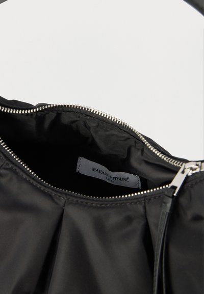 Μαύρη τσάντα με φερμουάρ ανοιγμένη, εμφανίζοντας την εσωτερική ετικέτα με το κείμενο «Maison Kitsuné Paris».