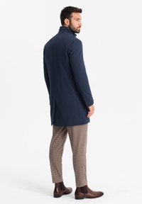 Cappotto navy con colletto alto, vestibilità slim e spacchi laterali, abbinato a pantaloni beige a motivo houndstooth e stivali Chelsea marroni.