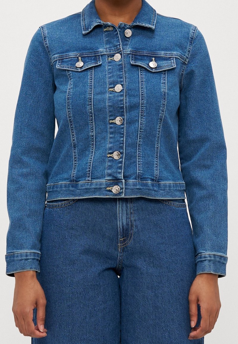 Denimjacka i medelblå, med en åtsittande design, synliga knappar, två bröstfickor och en nederkant som slutar vid midjan.