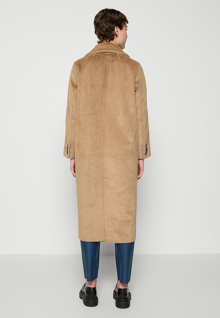 Brązowy płaszcz do kolan z miękkiego, teksturowanego materiału z klapami i zapinanymi mankietami, w zestawieniu z niebieskimi skórzanymi spodniami i czarnymi butami.