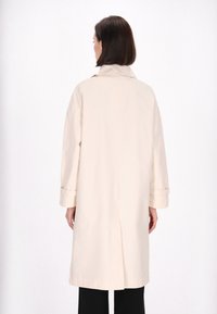 Beige trenchcoat med en afslappet pasform, stort revers og opslåede manchetter. Har en midtertransportventil for bevægelsesfrihed. Glat, letvægtsstof.