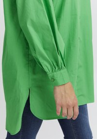 Green button-up shirt with long sleeves, gathered cuffs, and a curved hem. Made of smooth fabric with a casual fit.