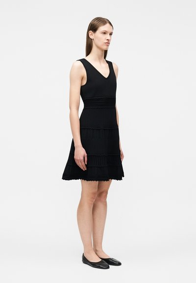 Femme debout portant une robe noire sans manches jusqu'aux genoux avec des bandes horizontales texturées et des chaussures plates noires, sur un fond uni.