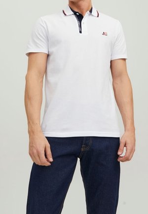 White short-sleeve polo shirt with a navy collar featuring red accents and a logo on the left chest, paired with dark blue jeans.