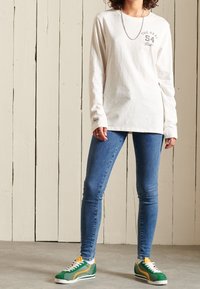 Långärmad krämfärgad t-shirt med grafiskt tryck, kombinerad med blå skinny jeans och gröna sneakers med gula accenter. Har en minimalistisk design.